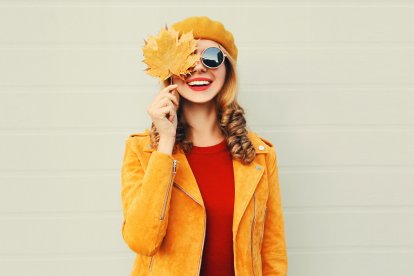 Autumn mood! happy smiling woman holding in her hands yellow maple leaves hiding her eye over gray wall background