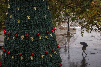 Una persona con paraguas caminando junto al árbol de Navidad.